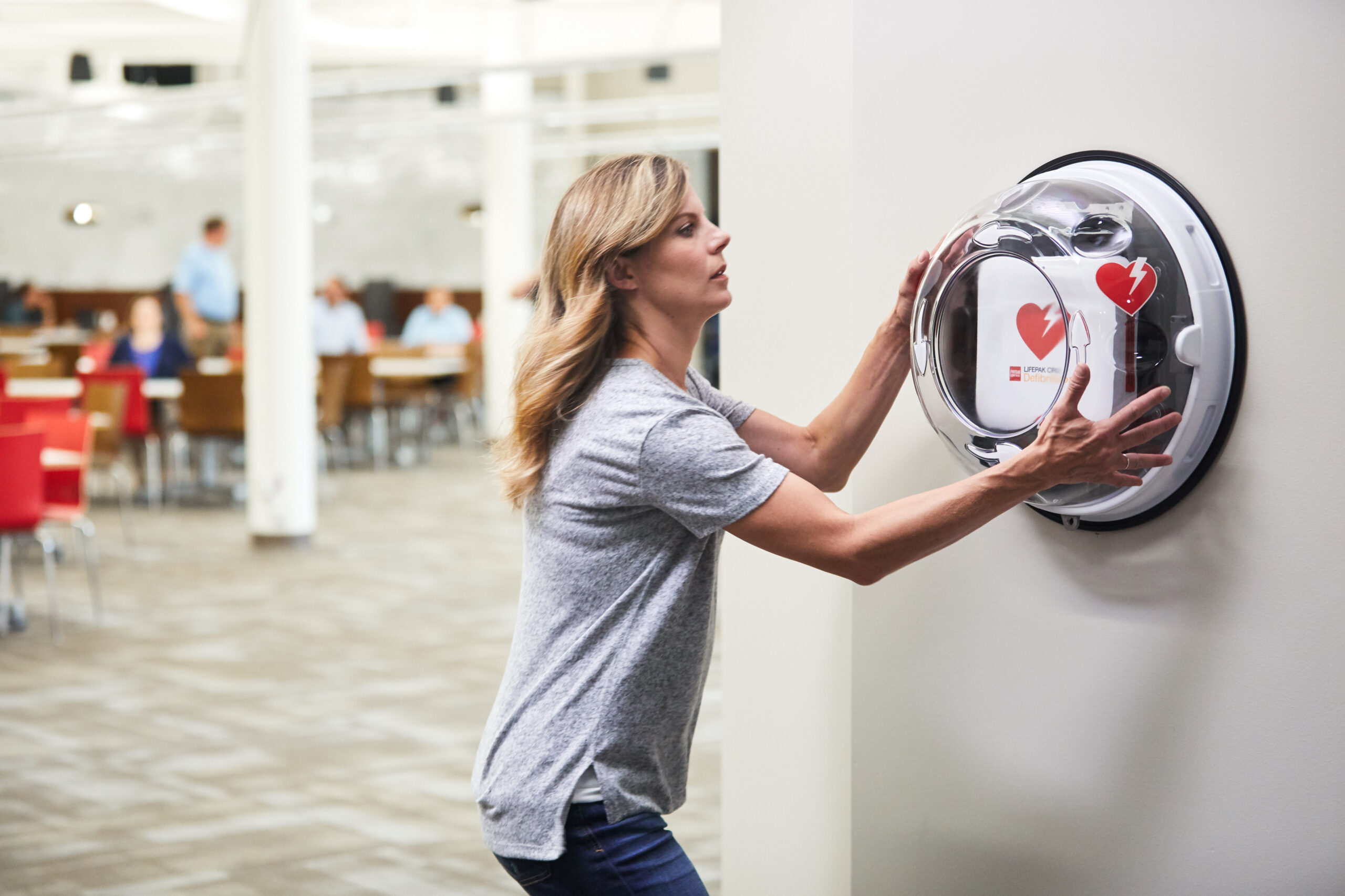 A woman reaches for an AED in a wall-mounted cabinet during a cardiac emergency