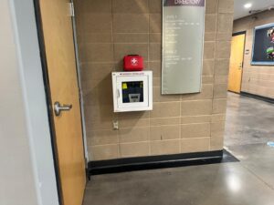 AED in a wall-mounted cabinet beside a building directory in a community center corridor, with a first aid kit resting on top of the cabinet