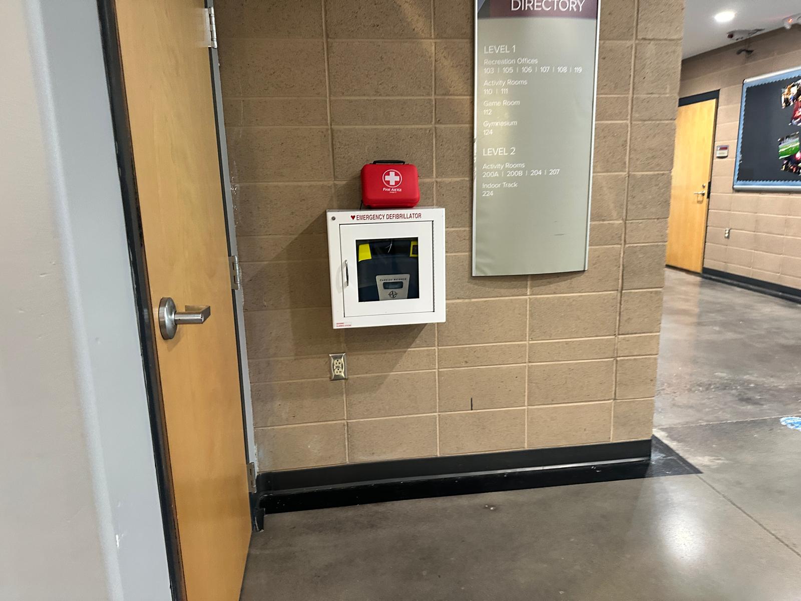 AED in a wall-mounted cabinet beside a building directory in a community center corridor, with a first aid kit resting on top of the cabinet